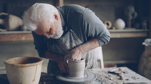 istock Experienced ceramist grey-haired bearded man is smoothing molded ceramic pot with wet sponge. Spinning throwing wheel, muddy work table and clayware are visible. 1436083253