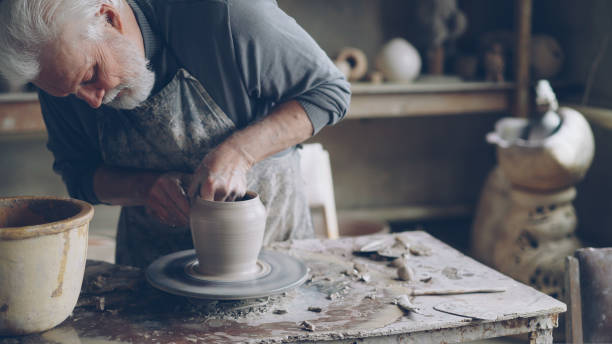 istock Experienced ceramist grey-haired bearded man is smoothing molded ceramic pot with wet sponge. Spinning throwing wheel, muddy work table and clayware are visible. 1436083318