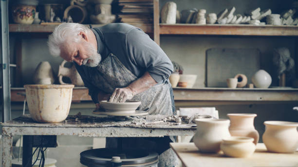 istock Experienced senior master is making low clay bowl on a pottery wheel in small workshop. Ceramics jigger, handmade clayware and potter's tools are visible. 1436083769