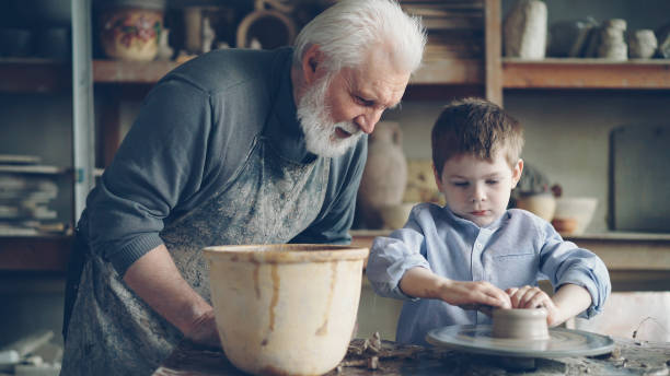 istock Concentrated young boy is molding clay into ceramic pot on spinning throwing wheel and his experienced grandfather is talking to him. Pottery and family tradition concept. 1436083880