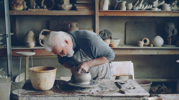 istock Skilled elderly potter is producing ceramic pot on turning wheel in workplace. Creation process, traditional pottery and interesting hobby concept. 1436085621