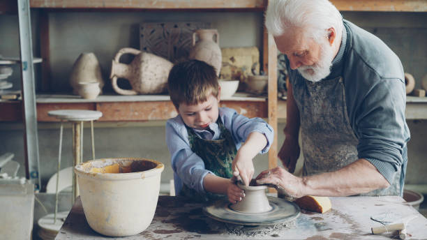 istock Concentrated little boy is learning to work with clay on throwing-wheel in pottery class in traditional workshop. His teacher senior experienced man is helping him. 1436086109