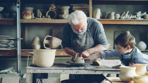 istock Cute boy is molding clay on throwing wheel while his caring grandfather is teaching and helping him, giving advices. Shelves with numerous ceramic pots in background. 1436086363