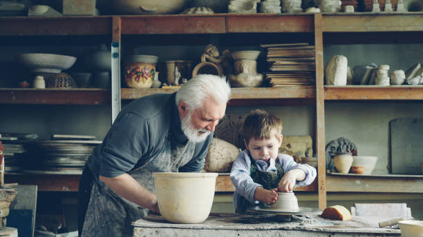 istock Curious boy is learning pottery from his experienced grandfather in small home studio. Child is forming clay to make pot on potter's wheel, his granddad is helping him. 1436086934