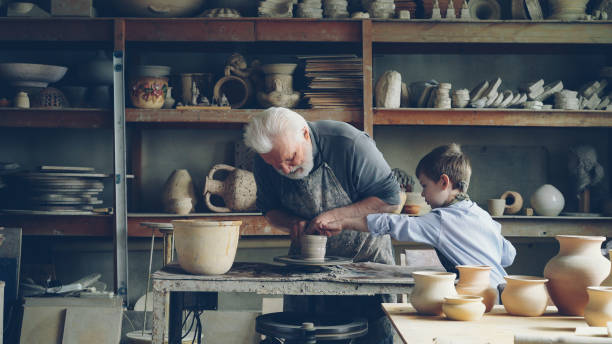 istock Professional male potter is working with clay on spinning throwing-wheel with his curious grandson helping him. Ceramic pots, vases and figures are visible. 1436086988