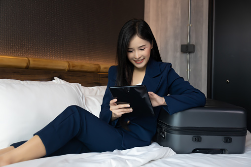 Business Technology Internet Hotel Vacation Concept Young businesswoman working from hotel room on business trip Asian woman sitting on bed and using digital tablet for chat to customer during travel