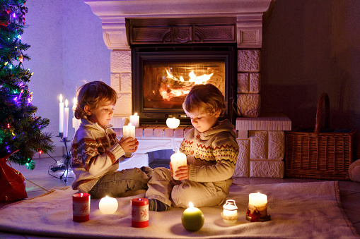 Two little children sitting by a fireplace at home on Christmas time. Cute toddler boys, blond twins playing together and lookinig on fire in chimney. Family celebrating xmas holiday