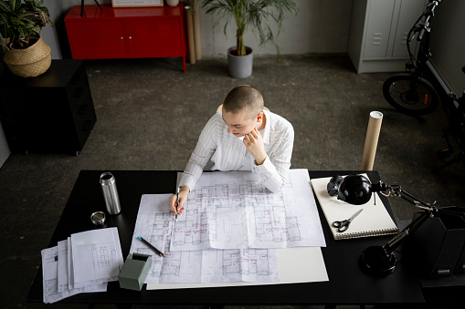Young bald lady analyzing blueprint at desk in office, working hard