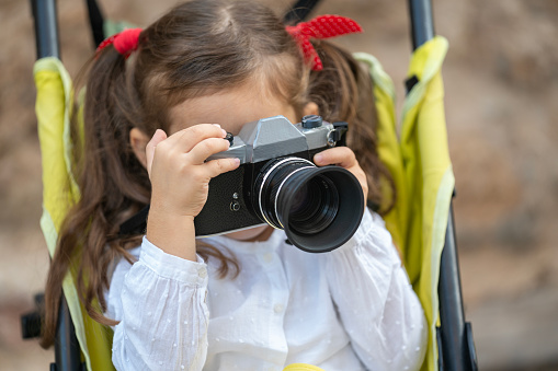 Photo of 4 years old girl photographing with analog camera. She is sitting on baby stroller in outdoor. Shot in outdoor with a full frame mirrorless camera.