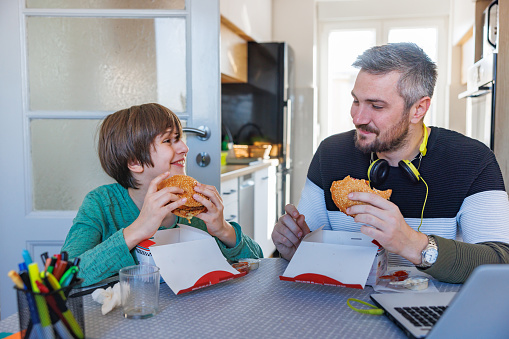 Father and son sitting at the table at home, eating burgers from takeout boxes and smiling at each other