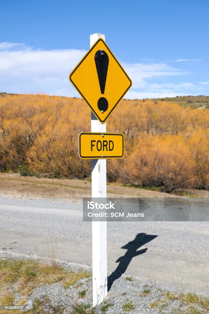 Ford Warning Post Road sign warning of a stream crossing ahead.  The sign has bullet holes from gun shots.  This image was taken near Lake Pukaki on a sunny afternoon in early Spring. Ford - Crossing Stock Photo