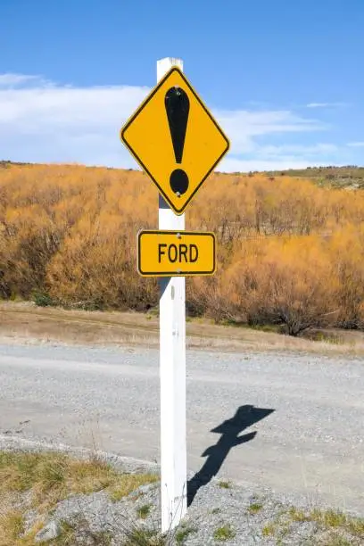 Road sign warning of a stream crossing ahead.  The sign has bullet holes from gun shots.  This image was taken near Lake Pukaki on a sunny afternoon in early Spring.