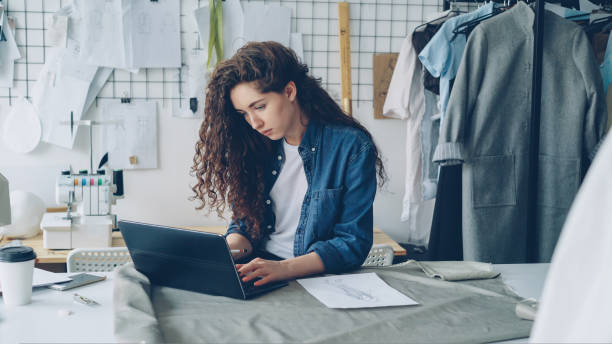 istock Self-employed seamstress is working with laptop sitting at table in modern light workshop. Woman is typing and watching screen and browsing for inspiration concept 1436094042