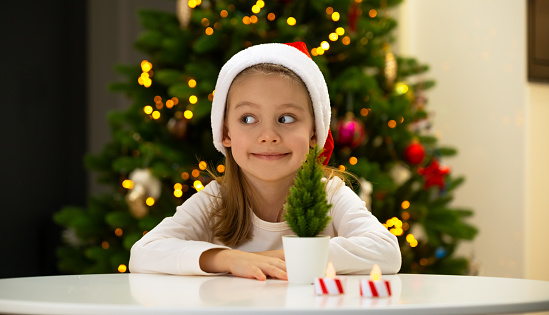 Little girl with dreamy look in red santa hat sits at table and smiles on background of shining Christmas tree. Happy child celebrates New year and christmas