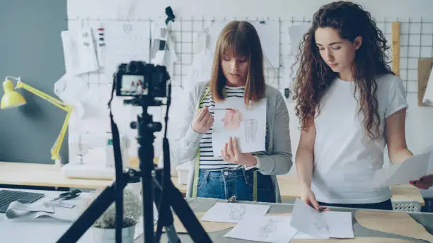 Photo of Young women fashion bloggers recording video blog about ladies'clothes on camera and talking to followers in modern studio. Many garment sketches are visible.