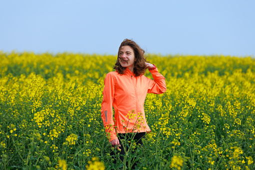 Young woman in orange windbreaker standing in agricultural field with yellow flowering plant on a clear windy day