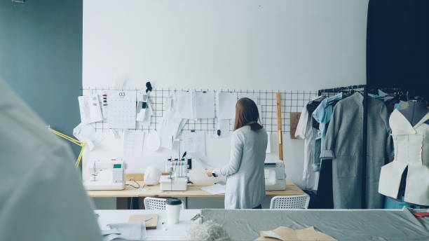 istock Back view of creative female tailor taking sketches from tailoring desk and hanging them on wall above table. Planning and creating new collection concept. 1436096084