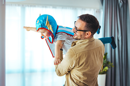 Dream big. Loving father helps his son fly like a superhero. Boy play fly with his dad at home. Cheerful familyare having fun together. Father holding son in superhero costume flying at home
