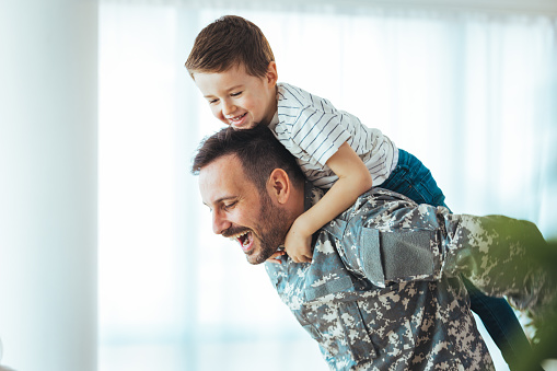 Happy reunion of one young soldier with family, five years old son and father having fun in the house during the beautiful sunny day. Soldier dad hugs his son after a time in military.