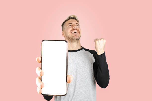 Caucasian young adult man smiling and celebrating while holding smart phone with white screen for copy space advertisement. Close up caption of mobile. Isolate pink background stock photo