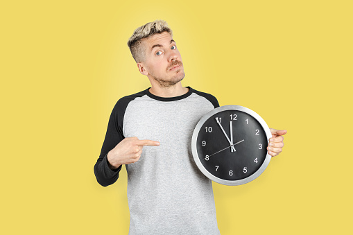 Caucasian man on casual clothing showing time on wall clock and pointing with finger with gesture of concern, Isolated yellow background