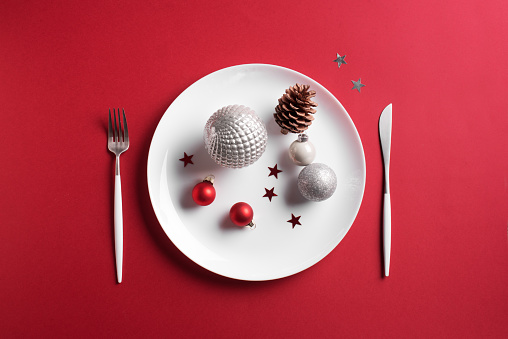 Beautiful Christmas Table Setting.  White plate, cutlery with and festive ornaments on red background, flat lay, copy space. Christmas dinner, party design, menu concept.
