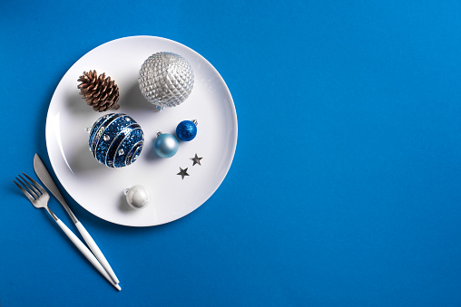 Creative Christmas Table Setting.  Festive ornaments and decor on white plate on blue background, top view, copy space. Christmas dinner, party design, menu concept.
