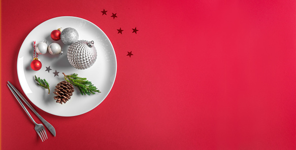 Beautiful Christmas Table Setting.  Festive silver baubles and pine cone on white plate on red background, creative flat lay, banner, copy space. Christmas dinner, party design, menu concept.