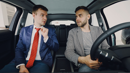 Young confident businessman is discussing new car with professional salesman sitting in front seats inside luxury automobile and holding wheel. Conversation and buying transport concept.