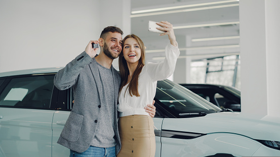 Attractive young couple is taking selfie with car keys in modern motor showroom after buying new automobile. They are looking at camera, posing, kissing and smiling.
