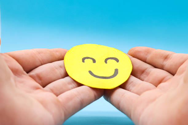 istock Hands holding smiley yellow paper face on a blue background. Happiness concept, positive thinking, mental health assessment, world mental health day concept, optimism 1436112236