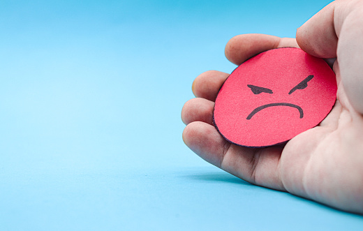 Hand holding a red paper angry face, on a blue background. Concept of aggressiveness, anger, hate, revenge, jealousy.