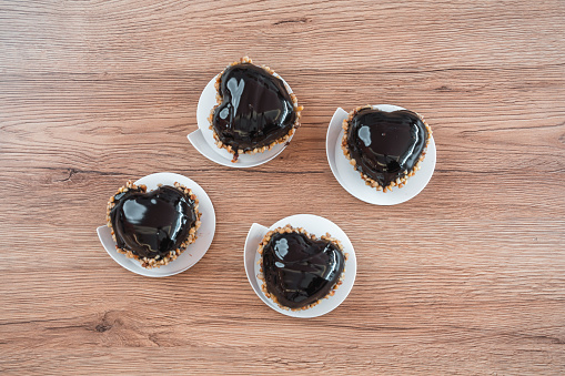 Bird's eye view of four small chocolate heart-shaped desserts, coated in glossy dark chocolate and dipped in crushed almonds. Sweets on little white serving trays standing on a wooden counter.