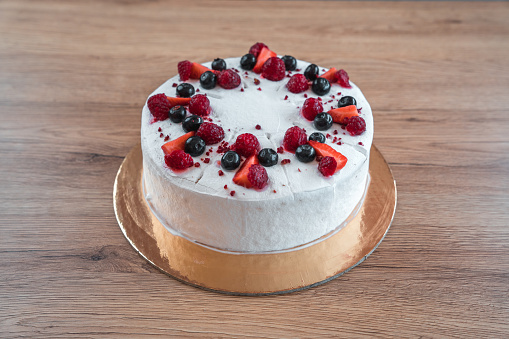 Whole, round cake prepared to be cut and served. Coated with white cream and topped with blueberries, raspberries and strawberries. High angle shot. Full length image.