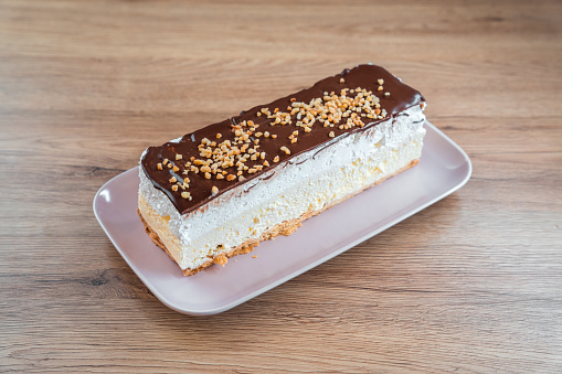 Full, uncut creamy dessert displayed on a white  serving plate. Layers made of custard cream and puff dough, top is decorated with chocolate and biscuit crumbs. High angle view. Wooden shelf under.