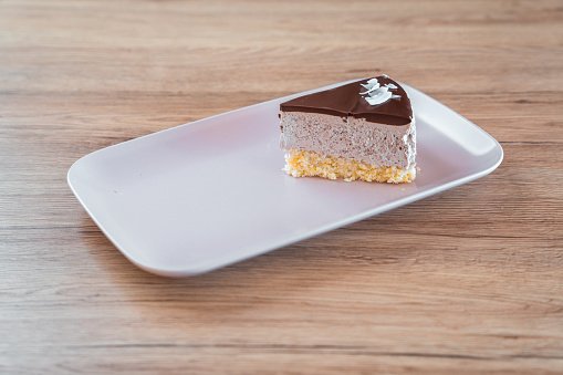 A slice of delicious cake on an elegant, white ceramic plate, being served at a patisserie. Layers of fruity cream and chocolate top layer visible from the side. High angle shot.