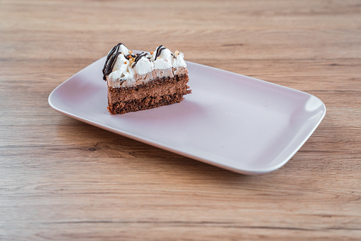 Professionally served slice of vegan cake, topped with abundance of whipped almond cream and dark chocolate. High angle view of cake on rectangular plate.