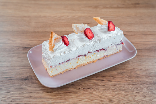 Long, white dessert displayed on a porcelain serving plate. Layers of custard, cream and puff dough visible on the sides, top is decorated with fresh strawberries and pieces of puff dough. High angle view.