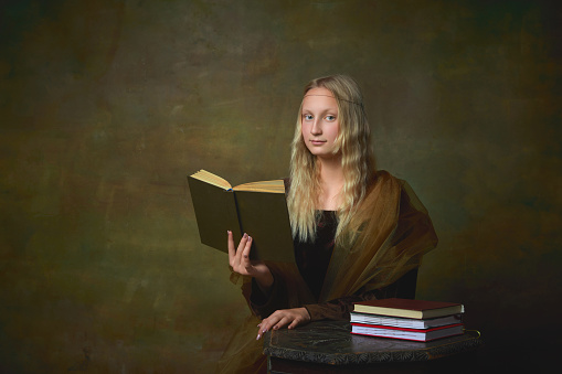 Beautiful young girl with long blonde hair wearing medieval dress reading book over dark vintage background. Retro style, fashion, comparison of eras concept. Beautiful female model like Mona Lisa