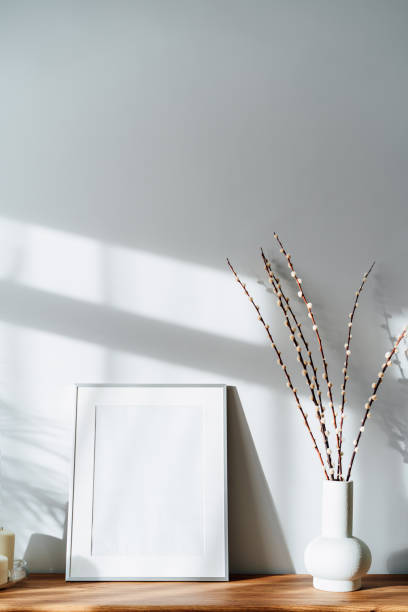 istock Modern minimalist style interior with white poster mockup, candles and blooming branches of the pussy willow in vase on wooden console under sunlight and shadows on a white gray wall. Selective focus. 1436124000