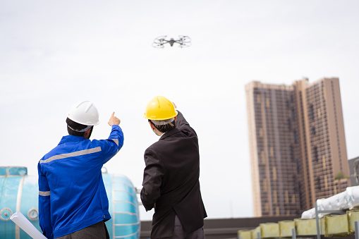 Professional Asian civil engineers using the unmanned aerial vehicle or UAV - drone for inspecting the building structure, 2 Asian foreman flying a drone to the building roof top area.