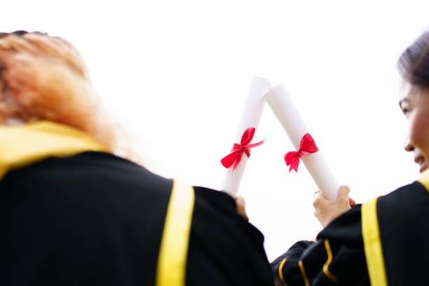 Happy Asian young women in the master degree gown showing a diploma in their hand close up. Portrait of confident college students in gown in the graduation ceremony.