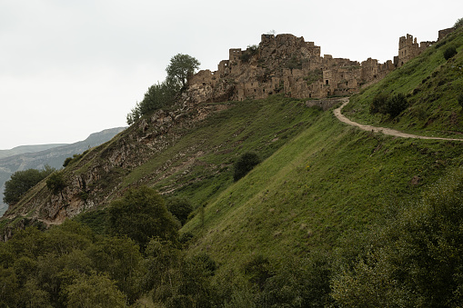 Ruin of old stone castle on peak of in mountain in Dagestan with road on green slope, majestic mountains landscape, canyon in highlands in overcast. Historic adventure, tourist trip.