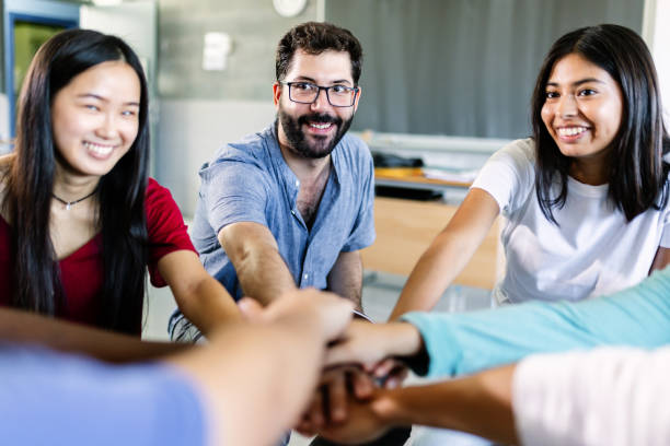 istock Young diverse group of students stacking hands with male teacher in classroom 1436127632