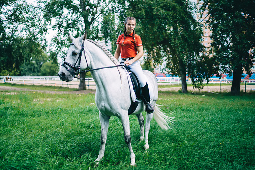 Portrait of professional female jockey practising horseback equitation riding and looking at camera during free time in country club, concept of equestrian sport and friendship with animal