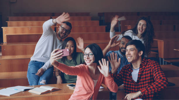 istock Joyful students are having fun recording video in classroom waving hands and smiling sitting at desks in lecture hall. Selfie, smartphone and education concept. 1436135917