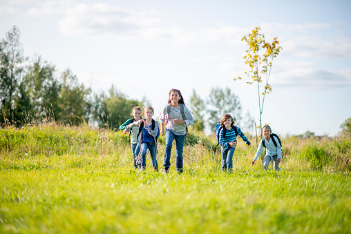 A small group of Elementary children run through a field as they make their way back to school for the first day.  They are each dressed casually and have their backpacks on as they run excitedly.
