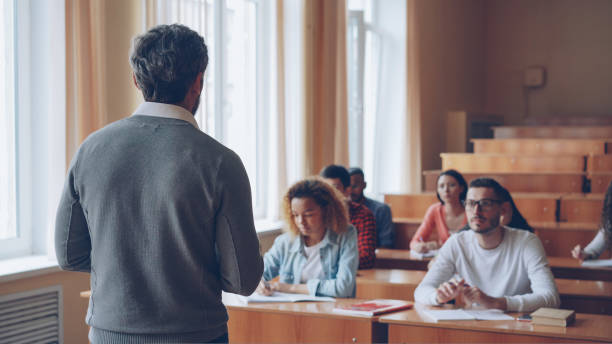 istock Bearded man professor is reading lecture talking and gesturing while students are listening and writing sitting at tables in spacious university classroom. 1436138301