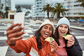 Selfie, pizza and friends with women taking a photograph on a phone outdoor in the city during winter. Fast food, happy and eating with a young female and friend outside in town on a urban background