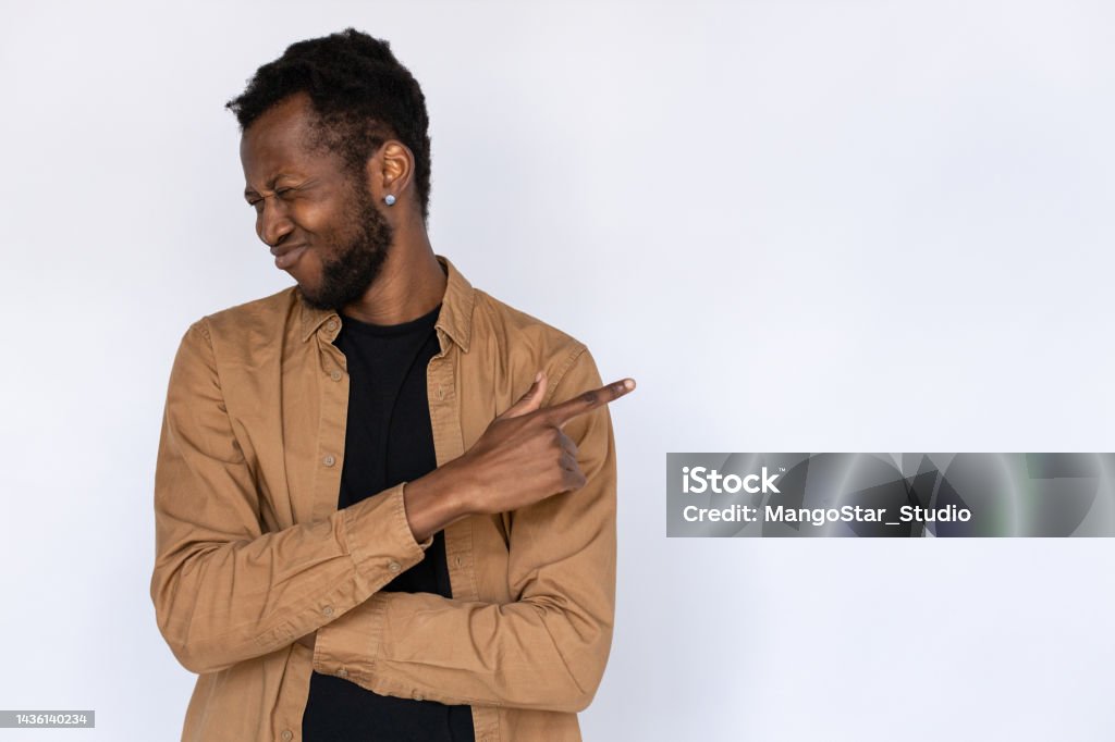 Disgusted man pointing aside Disgusted man pointing aside. Male African American model in black T-shirt and brown shirt pointing at something with disapproval, turning face away. Portrait, studio shot, advertising concept Men Stock Photo